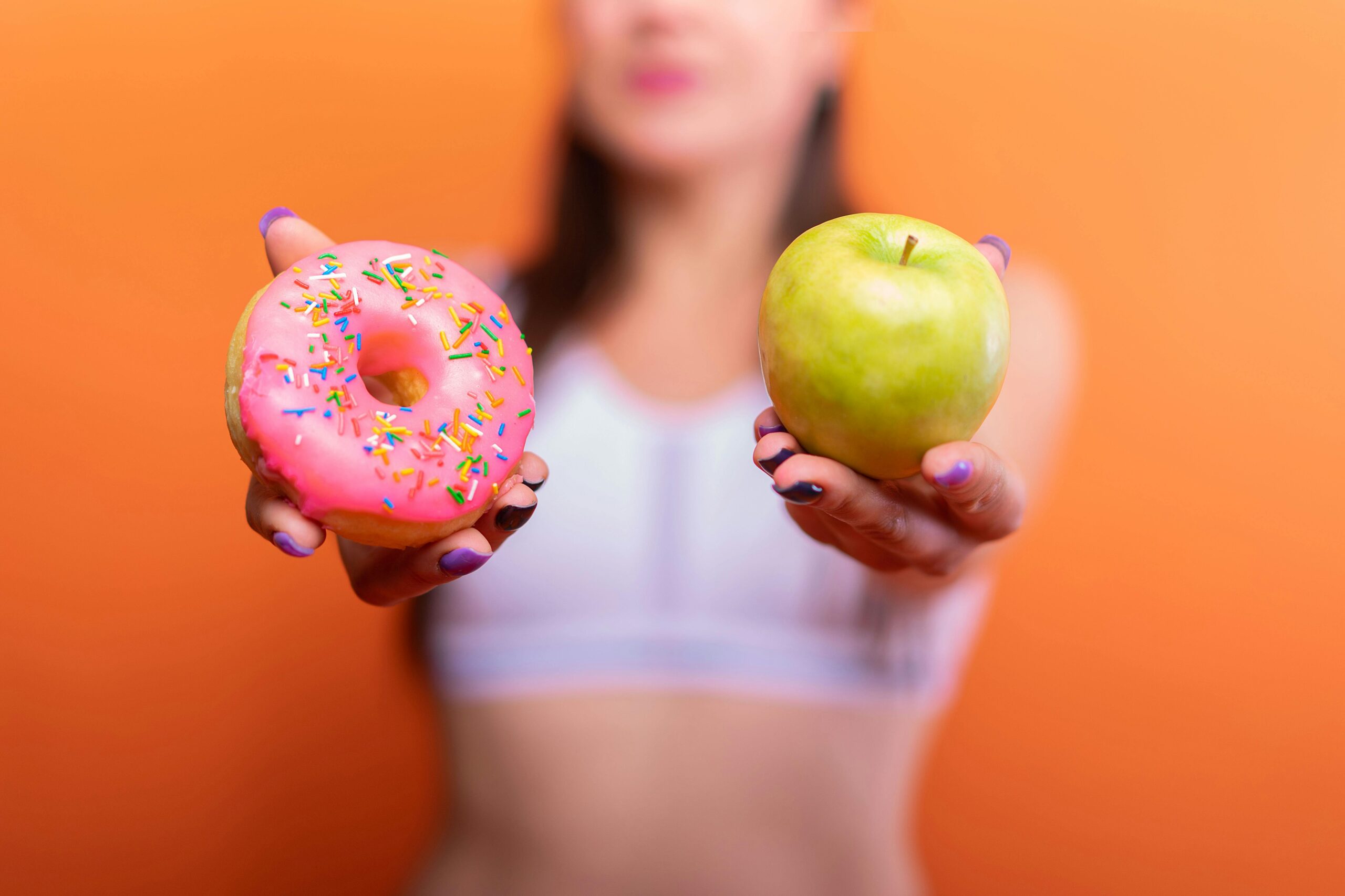 A woman holds a green apple and a pink donut with sprinkles against an orange background, emphasizing choice.