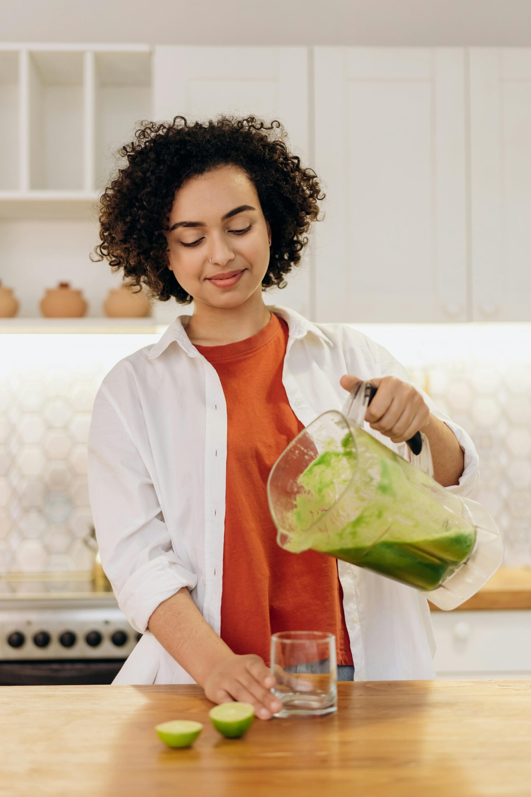 Woman in casual attire pouring a fresh green smoothie into a glass in a modern kitchen.